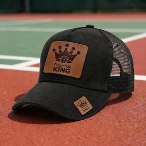May include: Black trucker hat with a mesh back and a curved brim. The hat features a brown leather patch with a crown and the words "Pickleball KING". A smaller patch with the same design is on the side.