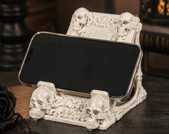 May include: A cream-colored phone stand featuring a detailed design of multiple human skulls and intricate bone-like textures. The stand holds a smartphone in a horizontal position, with two prominent skulls at the base and two at the top corners.