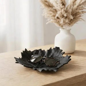 May include: A black leaf-shaped decorative tray holds a pair of sunglasses with dark lenses. A white vase with dried pampas grass sits in the background on a wooden surface. The tray is a stylish accent piece.