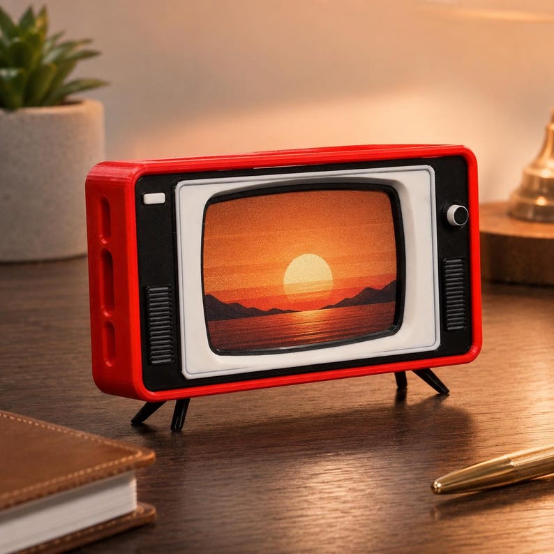 May include: A small, rectangular, red television-shaped decorative object with a white screen frame and black accents. The screen displays a printed image of a sunset over water with mountains in the background. It stands on four thin black legs.