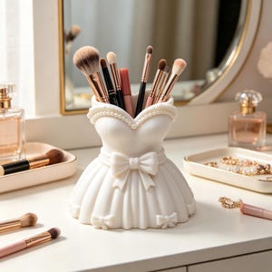 Könnte beinhalten: Ein weißer Make-up-Pinselhalter in Form eines Kleides mit ausgestelltem Rock, einer taillierten Taille mit Schleife und einem herzförmigen Ausschnitt, der mit einer Perlenkette verziert ist. Der Halter enthält verschiedene Pinsel mit braunen und schwarzen Borsten.