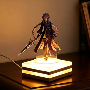 以下が含まれることがあります： 紫色の髪と暗い衣装のキャラクターのフィギュア。剣を持っています。フィギュアは、木製のフレームが付いた、光る正方形のベースの上に立っています。ベースは暖かく柔らかな光を発しています。