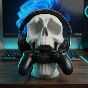 May include: A white skull-shaped headphone and controller stand. The stand holds a black gaming controller in its mouth and a pair of black headphones on its head. The stand is on a wooden surface.