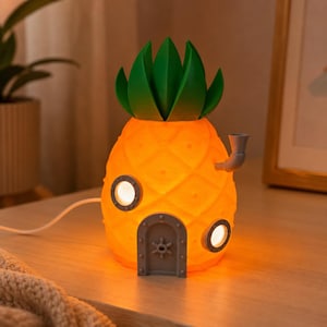 May include: A pineapple-shaped lamp with a bright orange glow, featuring a green leafy top, a grey arched door with a ship wheel design, two circular grey porthole windows, and a small grey chimney pipe, connected by a white power cord.