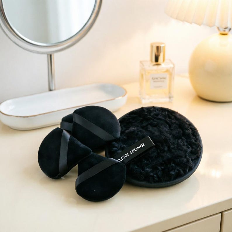 May include: A collection of black makeup applicators and a round, fluffy face cleaning pad. The applicators are in the shape of teardrops with black straps. The cleaning pad has a black strap that reads "CLEAN SPONGE".