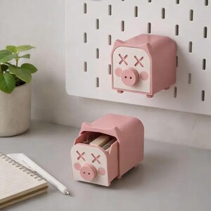 May include: Two pink pig-shaped storage drawers with white faces featuring X-shaped eyes and a circular snout. One drawer is mounted on a white pegboard, while the other sits on a surface with a small open compartment revealing contents.