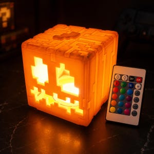 May include: A square, orange, pixelated jack-o'-lantern lamp with a glowing interior, featuring a carved face design. Beside the lamp sits a white rectangular remote control with multiple coloured buttons for adjusting light settings and colours.