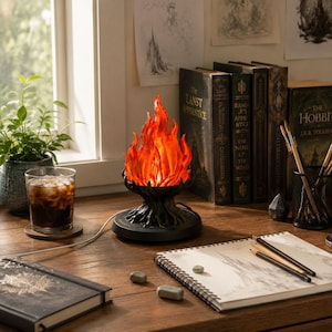 May include: A decorative lamp featuring a sculpted black base shaped like a tree stump, topped with a translucent orange and red flame-shaped light fixture. The lamp sits on a wooden desk next to a glass of dark liquid, books, and a sketchbook.