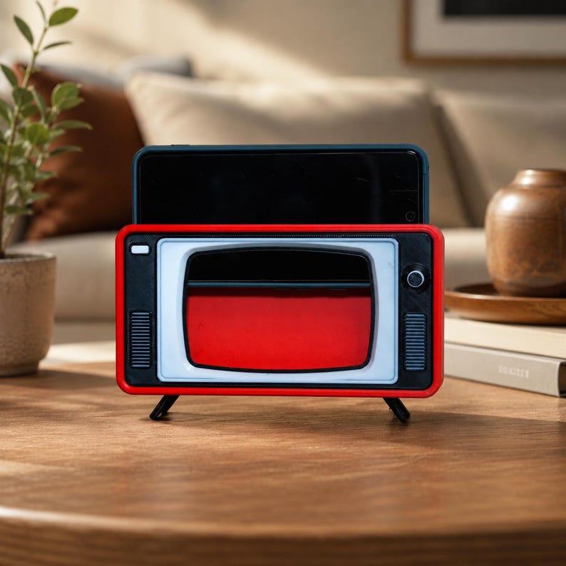 May include: A red and black smartphone stand designed to resemble a vintage television set. The stand features a rectangular frame with a white screen border, a small circular knob on the right side, and two black vertical speaker grilles on the front.