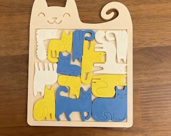 Puede incluir: Rompecabezas de madera con forma de gato con una cara sonriente y una cola rizada. Las piezas del rompecabezas son varias formas de gato en amarillo, azul y blanco. El rompecabezas está sobre una superficie de madera.