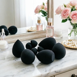 May include: A collection of black makeup sponges in various shapes and sizes, including teardrop and rounded forms, arranged on a marble countertop. The scene includes beauty products, a mirror, and a vase of pink roses, suggesting a beauty or cosmetic theme.