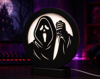 Puede incluir: Una lámpara circular negra que presenta la silueta recortada de una figura encapuchada con máscara y cuchillo. El diseño está retroiluminado con un suave resplandor blanco, creando una imagen de alto contraste. La lámpara tiene una base negra.