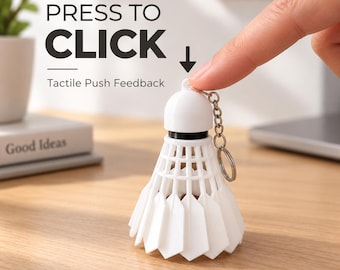 Puede incluir: Un llavero de volante de bádminton blanco con un mecanismo de botón pulsador. El texto PRESS TO CLICK y Tactile Push Feedback es visible sobre el objeto. El volante tiene una parte superior redondeada, un anillo negro y una falda.