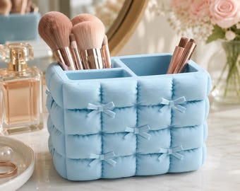 Puede incluir: Un soporte rectangular para brochas de maquillaje en color azul claro con textura acolchada y pequeños lazos decorativos en el frente. El organizador cuenta con varios compartimentos que contienen brochas con virolas de oro rosa.