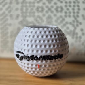 May include: White golf ball-shaped vase with dimples, featuring the "TaylorMade" logo in black. A small red "1" is visible below the logo. The vase sits on a wooden surface.