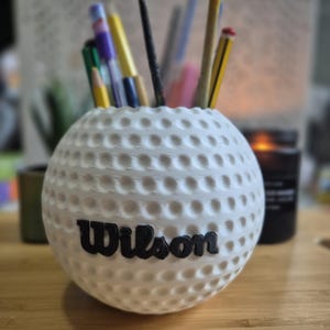 Peut inclure: Un pot à crayons blanc en forme de balle de golf avec le nom "Wilson" en lettres noires. Le pot contient divers stylos et crayons. Le design de la balle de golf présente des alvéoles sur sa surface.