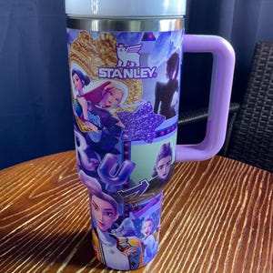 May include: A large, insulated tumbler with a purple handle and lid. The tumbler is decorated with a collage of cartoon characters, a purple star, and the "STANLEY" logo. The tumbler is sitting on a wooden table.