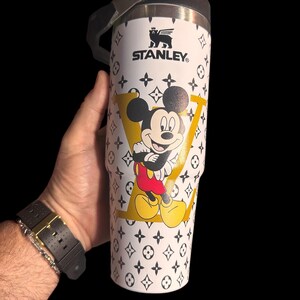 May include: White Stanley travel mug with a Mickey Mouse graphic inside a yellow "LV" design. The mug has a black handle and lid, and is decorated with a black and white pattern. The Stanley logo is visible.