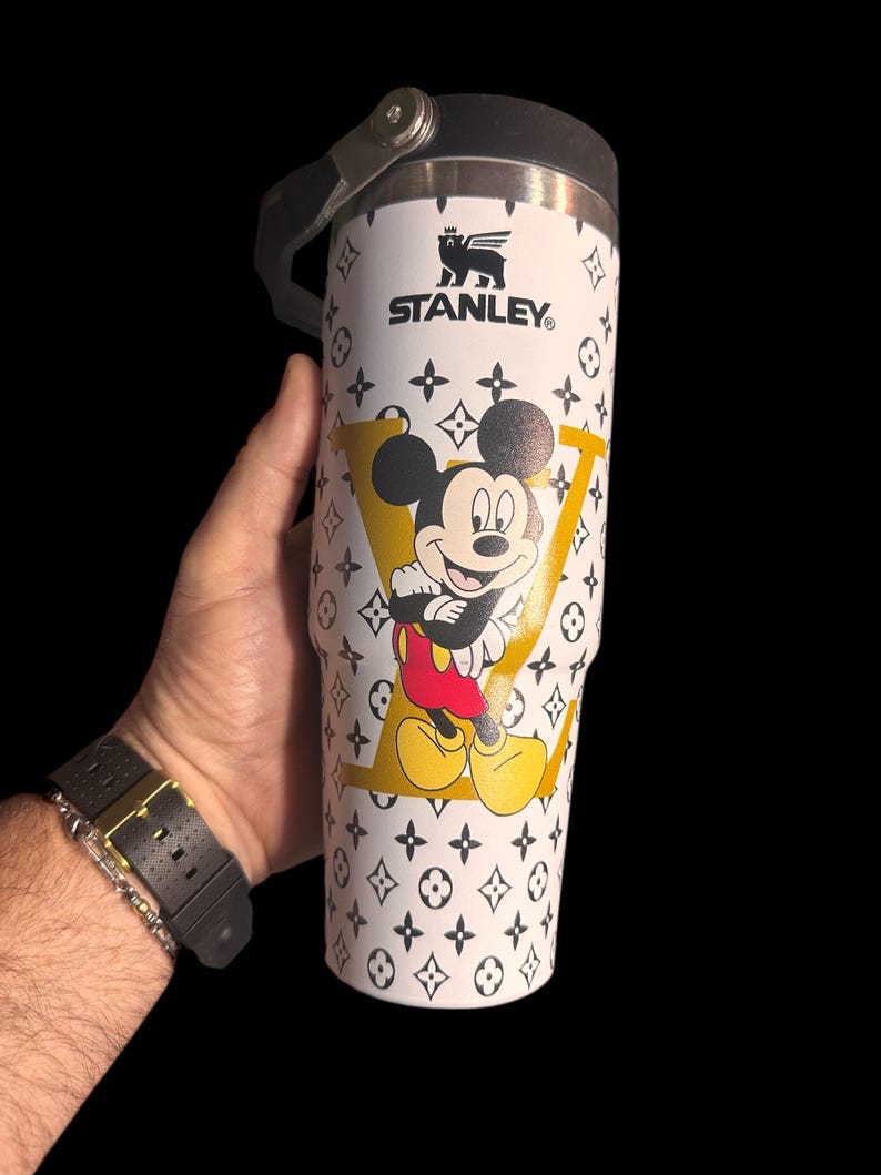 May include: White Stanley travel mug with a Mickey Mouse graphic inside a yellow "LV" design. The mug has a black handle and lid, and is decorated with a black and white pattern. The Stanley logo is visible.