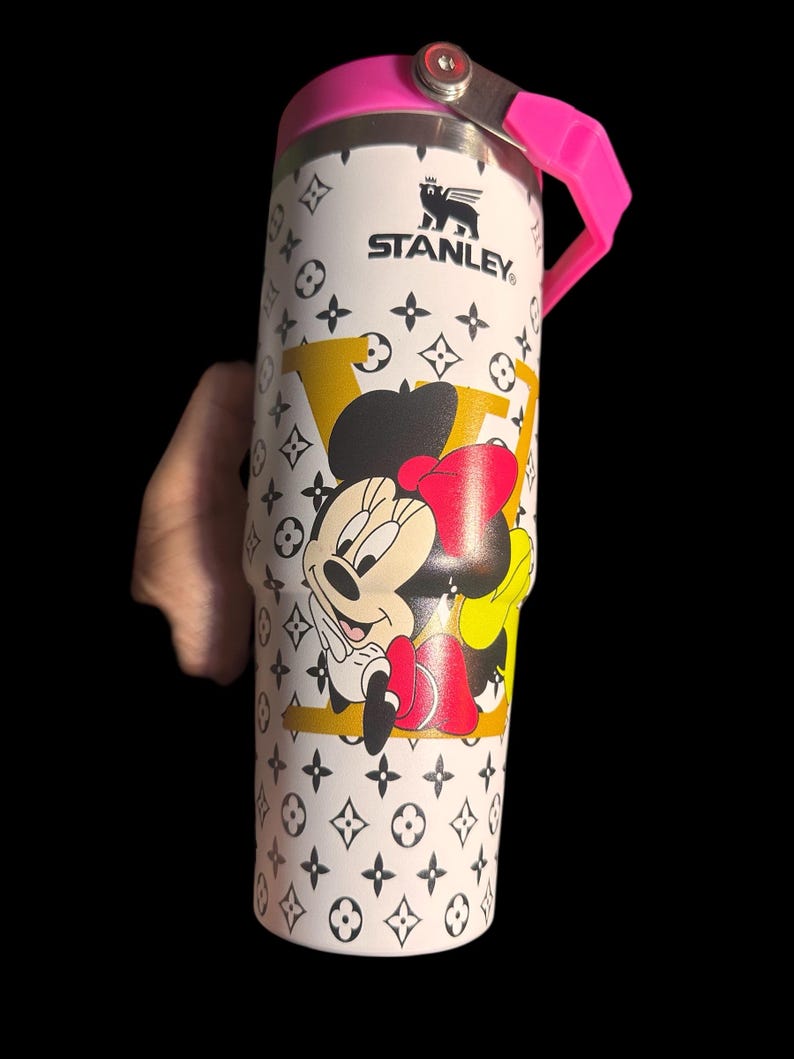 May include: White Stanley tumbler with a pink lid and handle, featuring a Minnie Mouse graphic and a repeating black and white pattern. The tumbler has the Stanley logo and a yellow and black design.