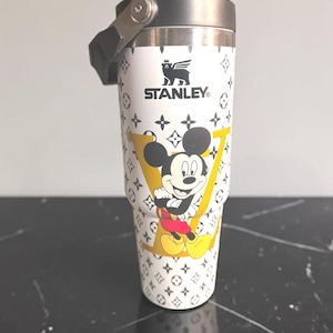 May include: White Stanley travel mug with a black handle and lid. The mug has a repeating pattern of black symbols and a large yellow "V" with Mickey Mouse. The Stanley logo is printed in black.