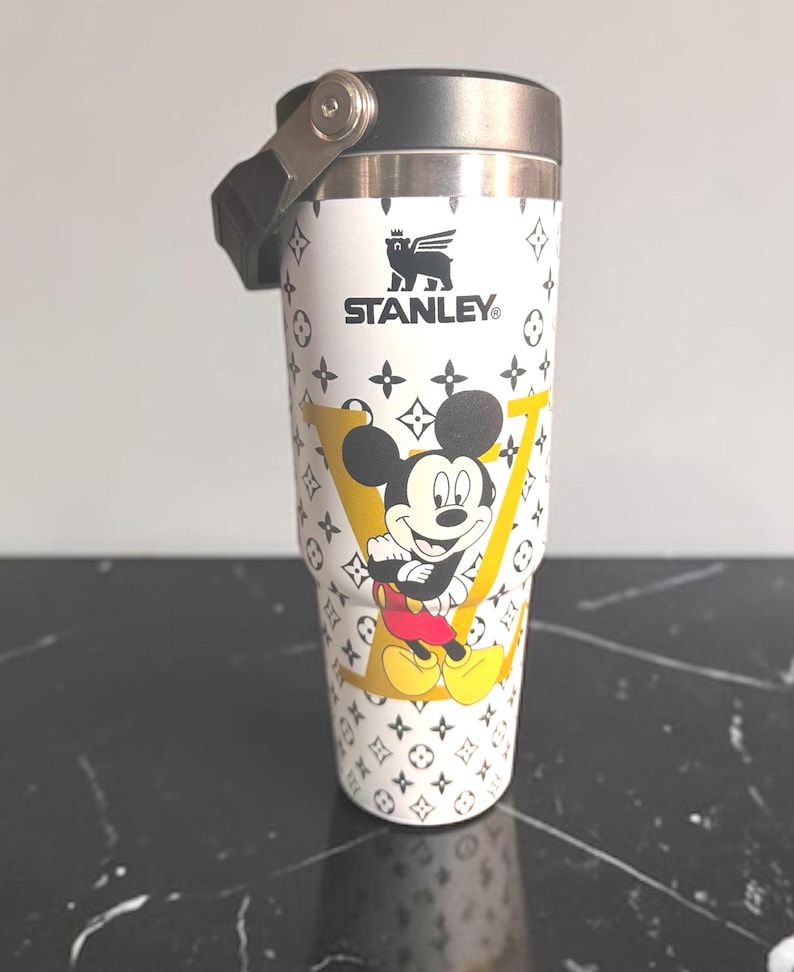 May include: White Stanley travel mug with a black handle and lid. The mug has a repeating pattern of black symbols and a large yellow "V" with Mickey Mouse. The Stanley logo is printed in black.