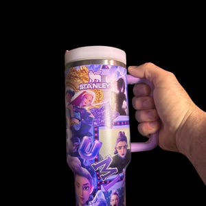 Puede incluir: Un vaso Stanley de acero inoxidable con asa y tapa moradas. El vaso est&aacute; cubierto con un collage de im&aacute;genes con la palabra "UMI" en letras moradas. El logotipo de Stanley es visible.
