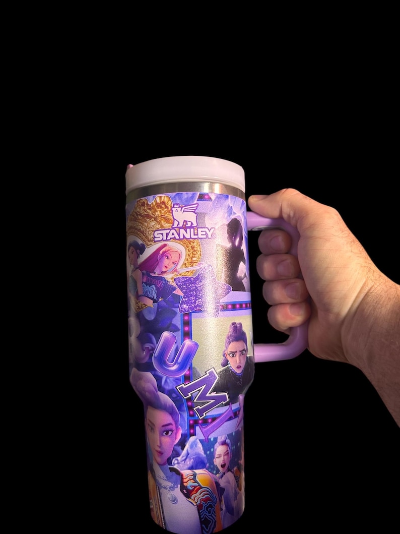 Puede incluir: Un vaso Stanley de acero inoxidable con asa y tapa moradas. El vaso est&aacute; cubierto con un collage de im&aacute;genes con la palabra "UMI" en letras moradas. El logotipo de Stanley es visible.