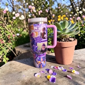 Puede incluir: Un vaso Stanley de acero inoxidable con tapa blanca y asa rosa. El vaso est&aacute; decorado con un collage de personajes de dibujos animados en tonos morados, azules y amarillos. La palabra "Stanley" es visible.