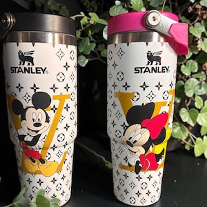 May include: Two Stanley travel mugs with Mickey and Minnie Mouse illustrations. The mugs have a white background with a repeating black pattern and a large yellow "V". One mug has a black lid, the other a pink lid. Each mug is approximately 20 cm tall.