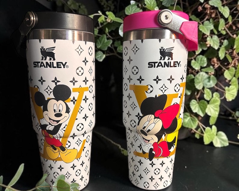 May include: Two Stanley travel mugs with Mickey and Minnie Mouse illustrations. The mugs have a white background with a repeating black pattern and a large yellow "V". One mug has a black lid, the other a pink lid. Each mug is approximately 20 cm tall.