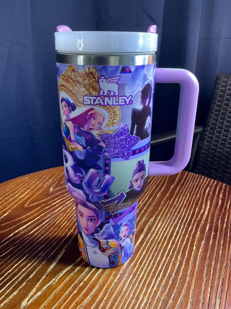 May include: A large, insulated tumbler with a purple handle and lid. The tumbler is decorated with a collage of cartoon characters, a purple star, and the "STANLEY" logo. The tumbler is sitting on a wooden table.