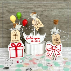May include: A selection of birthday gifts with glass vials, cork stoppers, and wooden tags. The gifts are decorated with balloons, gift boxes, and the text "Happy Birthday" and "Geldregen für dich". The overall theme is celebratory.