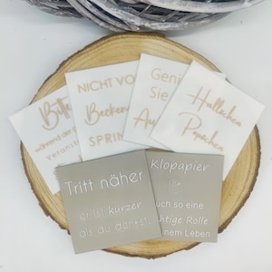 Könnte beinhalten: Eine Sammlung quadratischer, dekorativer Schilder mit deutschem Text in verschiedenen Schriftarten und Farben. Die Schilder sind auf einer Holzscheibe angeordnet. Der Text enthält Sätze wie "Tritt näher" und "Klopapier".