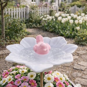 May include: A white, flower-shaped bird bath with a pink bird floating in the water. The bird bath is supported by a wooden pole and surrounded by colourful flowers. The background features a garden with a white picket fence and a house.