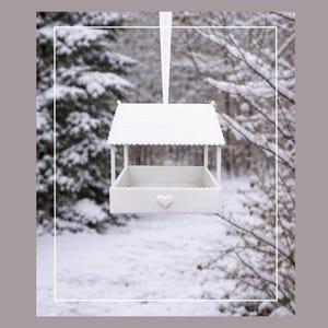 May include: A white bird feeder with a heart-shaped detail, hanging from a white ribbon. The feeder has a roof and a tray-like base. The background shows a snowy forest scene.