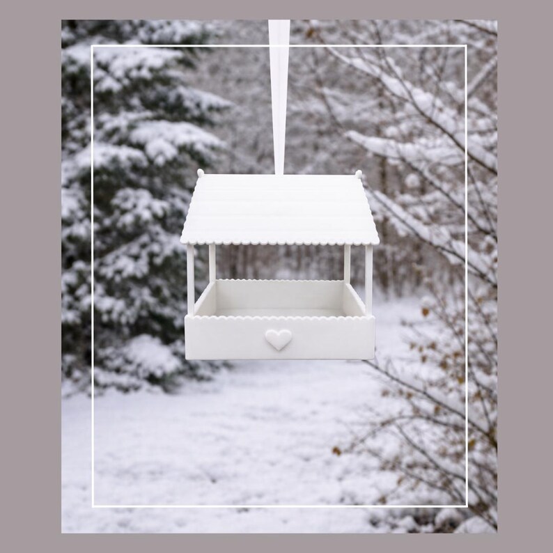 May include: A white bird feeder with a heart-shaped detail, hanging from a white ribbon. The feeder has a roof and a tray-like base. The background shows a snowy forest scene.