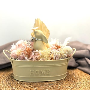 Op de afbeelding: Een decoratief arrangement in een crèmekleurige metalen container met het woord "HOME" in reliëf. Het bevat een keramieken konijnenfiguur, gedroogde bloemen en een klein belletje. De bloemen zijn in tinten roze, oranje en crème.