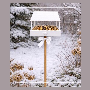May include: A white wooden bird feeder with a roof, filled with birdseed, and a heart-shaped detail. It is mounted on a wooden pole and adorned with a white bow. The background shows a snowy winter scene.