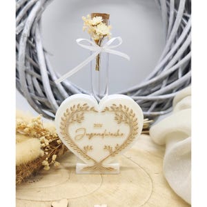 Può includere: Una candela a forma di cuore, bianco sporco, con la scritta dorata "Zum Jugendweihe", incorniciata da un disegno a foglie dorate. Un piccolo flacone di vetro con fiori secchi e un nastro bianco è posto sopra. Sfondo con una corona bianca e cuori di legno.