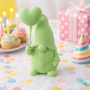 Könnte beinhalten: Eine hellgrüne Zwergfigur, die einen herzförmigen Ballon hält. Der Zwerg steht auf einer weißen Oberfläche mit bunten Konfetti, Cupcakes, einer Partyhütchen und einem verpackten Geschenk im Hintergrund. Die Szene deutet auf eine Geburtstagsfeier hin.