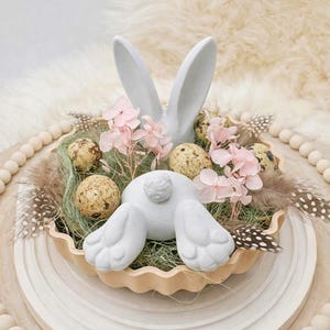 May include: An Easter-themed centerpiece featuring a white bunny with visible ears and hindquarters. The arrangement includes speckled eggs, pink flowers, and decorative feathers, all nestled in a scalloped dish on a wooden base. The overall color palette is soft and pastel.