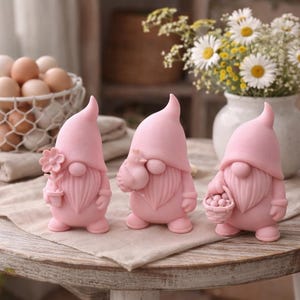 Puede incluir: Tres figuras de gnomos rosas con sombreros puntiagudos y barbas se exhiben sobre una mesa de madera. Uno sostiene una flor, otro un saco y el tercero una cesta de bayas. Una cesta de huevos y un jarrón de flores están en el fondo.