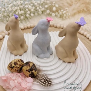 Könnte beinhalten: Drei Hasenfiguren in Beige und Grau, jede mit einem Schmetterling auf der Nase, sitzen auf einem weißen, geschichteten Tablett. Zwei gefleckte Wachteleier, eine rosa Blume und eine gemusterte Feder sind ebenfalls zu sehen. Das Bild hat eine weiche, pastellfarbene Ästhetik.