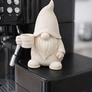 Könnte beinhalten: Cremefarbene Zwergfigur, die eine kleine Tasse mit der Aufschrift "COFFEE LOVER" hält. Der Zwerg hat einen langen weißen Bart und eine spitze Mütze. Er sitzt auf einer schwarzen Oberfläche, im Hintergrund eine schwarze Espressomaschine.