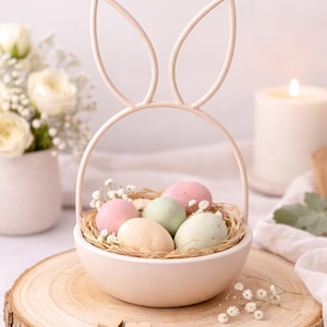 May include: A pastel pink Easter basket with bunny ear handles, filled with speckled eggs in pink, green, and beige. The basket sits on a wooden slice with two small wooden hearts, with flowers and a candle in the background.