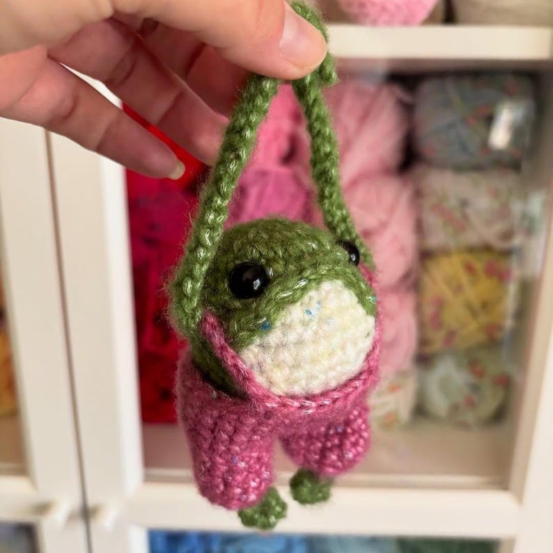 May include: A handmade crochet frog with a green body, white belly, and pink pants. The frog has large black eyes and is suspended by a green crocheted strap. The frog is a whimsical, handcrafted item.