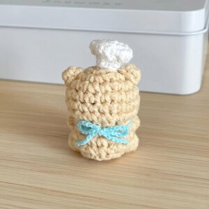 May include: A crocheted bear figurine wearing a white chef's hat and a light blue bow. The bear is tan and has a handmade appearance. The figurine is a decorative item.