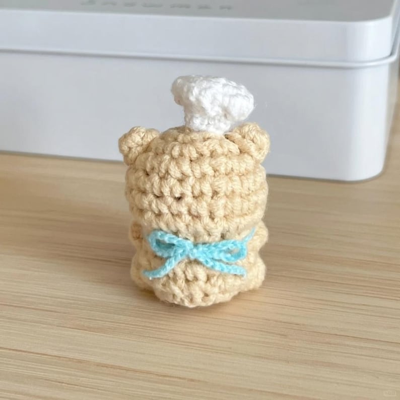 May include: A crocheted bear figurine wearing a white chef's hat and a light blue bow. The bear is tan and has a handmade appearance. The figurine is a decorative item.