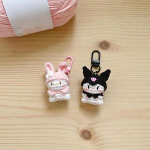 May include: Two crocheted keychains: a pink bunny with a pink clip and a black and white character with a black clip. The bunny has a pink hat and bow. The other character has black ears and a black hat. A ball of pink yarn is in the background.